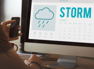 A person holding a phone while looking at a computer screen with a storm weather forecast.