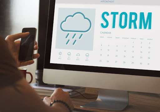 A person holding a phone while looking at a computer screen with a storm weather forecast.